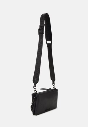 Sac bandoulière en cuir noir avec une large sangle réglable présentant un texte embossé et une fermeture éclair sur un fond uni.