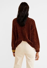 Person von hinten gesehen, der einen braunen strukturierten Pullover mit hochgekrempelten Ärmeln und cremefarbenen Hosen vor einem einfarbigen Hintergrund trägt.