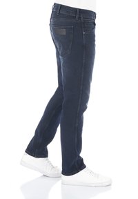 Jeans de mezclilla azul oscuro con un corte ajustado, que cuentan con un parche de cuero en el bolsillo trasero y costuras en contraste a lo largo de los lados. Zapatillas blancas.