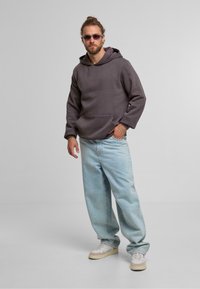 Felpa grigia con cappuccio e tasca frontale, abbinata a jeans larghi blu chiaro e sneakers bianche. Gli occhiali da sole completano l'outfit.