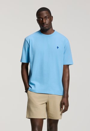 Man wearing light blue short-sleeve t-shirt with small dark blue logo and beige shorts, standing with one hand in pocket on gray background.