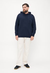 Felpa blu navy con tasca a marsupio e cappuccio con lacci, abbinata a pantaloni color crema e sneakers nere. Texture liscia, design casual.