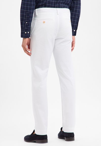 Weiße Baumwollhosen mit geradem Schnitt, ausgestattet mit Gesäßtaschen und Knopfdetails. Das Modell kombiniert sie mit dunklen Schuhen.