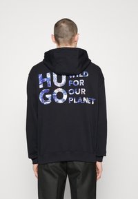 Sweat à capuche noir avec une capuche arrondie, présentant un grand imprimé de texte graphique au dos en bleu et blanc, portant la mention "HUGO WILD FOR OUR PLANET."