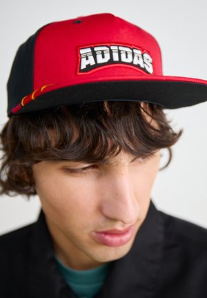 Casquette rouge et noire à visière plate, avec un logo "ADIDAS" brodé à l'avant, fabriquée en tissu structuré avec une texture lisse.