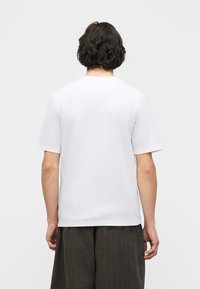 Άνδρας με σκούρα μαλλιά μέχρι τους ώμους, που φορεί απλό λευκό κοντομάνικο μπλουζάκι και σκούρο χαλαρό παντελόνι, στέκεται με την πλάτη προς τα έξω σε λευκό φόντο.