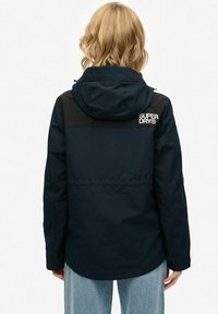 Granatowa wodoodporna kurtka z czarnym panelem na ramieniu i kapturem. Posiada logo z tyłu oraz wciągniętą talię, co nadaje dopasowany wygląd.