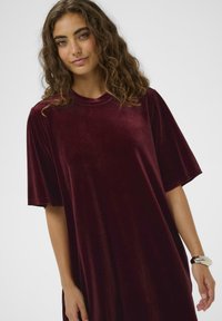 Rochie din catifea burgundy cu mâneci scurte și decolteu rotund, având o textură netedă și o croială lejeră. Design minimalist.