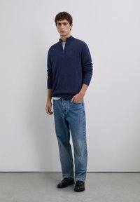 Man standing wearing a navy blue quarter-zip sweater, light gray t-shirt, blue jeans, and black leather shoes against a plain gray background.