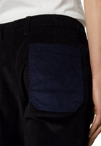 Pantalon en velours côtelé noir avec une grande poche arrière rectangulaire bleu marine foncé, porté par une personne vêtue d'une chemise blanche, main reposant le long du corps.