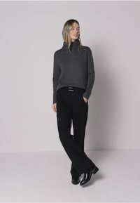 Pull à col roulé gris en tissu doux, associé à un pantalon noir ample et des bottes noires brillantes, posé devant un fond neutre.