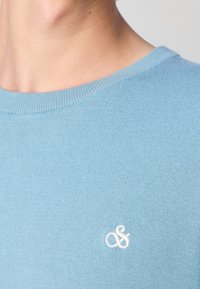Gros plan d'une personne portant un pull en maille bleu clair avec un petit logo blanc brodé près de l'épaule.
