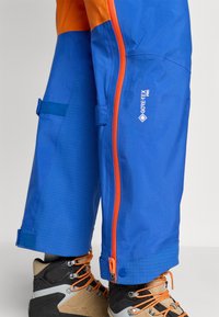 Pantaloni GORE-TEX Pro blu e arancioni, con chiusura a zip, orli regolabili, dettagli a motivo a griglia e texture del tessuto resistente.