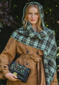 Brown suede trench coat with a tied waist, paired with a green plaid shawl. Holding a black clutch with colorful floral embroidery.