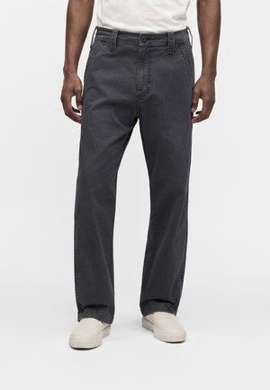 Mann trägt lockere dunkelgraue Hose, weiße Slipper und ein weißes Hemd, steht vor neutralem Hintergrund.