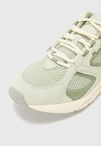 Încălțăminte sport de culoare verde deschis, realizată din materiale din plasă și piele întoarsă, cu o suprafață texturată, șireturi plate și o talpă căptușită. Accente notabile de culoare verde măsliniu.