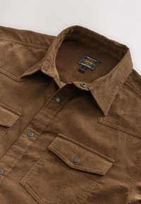 Chemise en velours côtelé marron à boutons, col pointu, deux poches avant à rabat avec boutons-pression, et une étiquette de marque visible à l'intérieur du col.