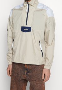 Beige vindjacka med vita axelapplikationer, framfickor med dragkedja, elastiska ärmslut och dragsko i nederkant. Columbia-logotyp på bröstet. Strukturerad tyg.