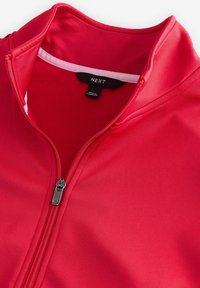Col montant de veste rouge à fermeture éclair montrant une fermeture argentée et une étiquette de marque noire "NEXT", avec une bordure intérieure blanche indiquant "ACTIVE".