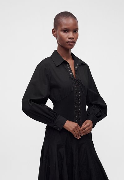 Femme aux cheveux courts portant une robe noire à manches longues avec un laçage devant et des poignets boutonnés, debout devant un fond uni.
