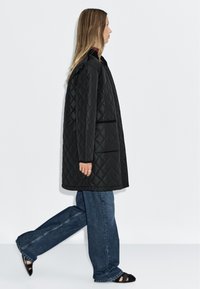 Schwarze gesteppte Jacke mit geradem Schnitt, langen Ärmeln und Seitentaschen, kombiniert mit weit geschnittenen blauen Jeans und schwarzen Sandalen.