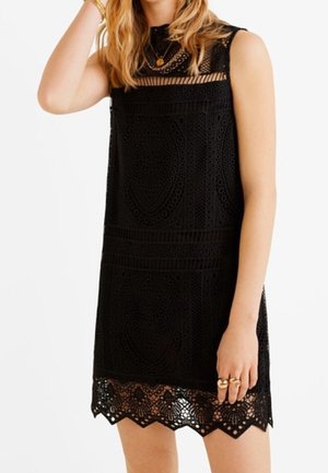 Robe mini sans manches en dentelle noire avec des motifs circulaires complexes et un ourlet festonné, portée avec des bagues et un collier dorés.