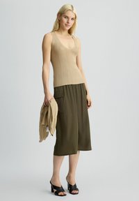 Canotta beige a coste, culotte verde oliva e sandali con tacco neri. La modella tiene un maglione beige coordinato, su uno sfondo chiaro.