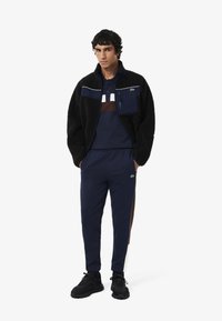 Czarna polarowa kurtka z zamkami, granatowy sweatshirt nałożony pod spód, granatowe dresy z brązowymi i białymi paskami po bokach oraz czarne sneakersy.