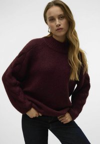 Burgunderfarbener gestrickter Pullover mit hohem Kragen und Kabelstrickärmeln, in lockerer Passform und weichem Material. Kombiniert mit dunklen Jeans.