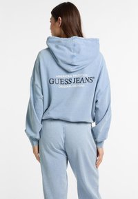 Zila kokvilnas kapucēta džempera ar lielu kapuci un pārāk lielu piegriezumu. Aizmugurē ir melns "GUESS JEANS" logo. Gluda, ikdienišķa auduma. Redzami saskaņoti bikses.