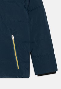 Sezione di giacca imbottita blu navy con una tasca con zip gialla e teal, tessuto texture con cuciture orizzontali e dettaglio nero sui polsini.