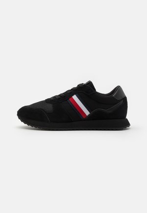 Zapatilla baja negra con suela negra y rayas laterales en rojo, blanco y azul, mostrada de perfil sobre un fondo blanco.