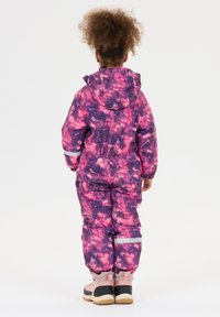Barn står med ryggen til iført en lilas og pink mønstret snedragt med reflekterende striber og pink vinterstøvler på en hvid baggrund.