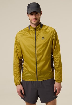 Hombre con chaqueta mostaza con cremallera y paneles laterales negros, pantalones cortos gris oscuro y gorra gris oscuro, de pie frente a un fondo neutro.