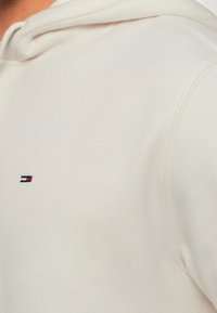 Mand iført en hvid hættetrøje med et lille, rektangulært logo i rød, hvid og marineblå broderet på brystet.
