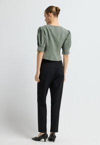 Frau mit einem Dutt, die eine grüne Bluse mit kurzen puffärmel und schwarze maßgeschneiderte Hosen sowie niedrige schwarze Absätze trägt, steht mit dem Rücken zur Kamera.