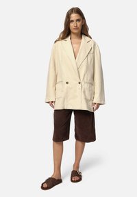 Beige dobbeltradet blazer med notch-lapper, to forlommer, parret med brune fløjlsshorts og brune fuzzy sandaler.
