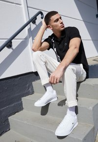 Černé polo tričko s bílou grafikou, krémové kalhoty a bílé tenisky. Model sedí na schodech s mírným záhybem na kalhotách.