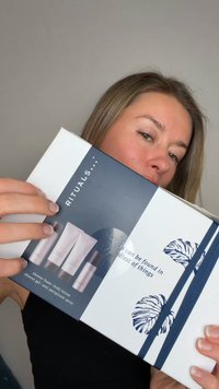 Hvit eske med mørkeblå aksenter og bånd, med Rituals-logo, som viser hår- og kroppspleieprodukter i myke, pastellfargede emballasjer.