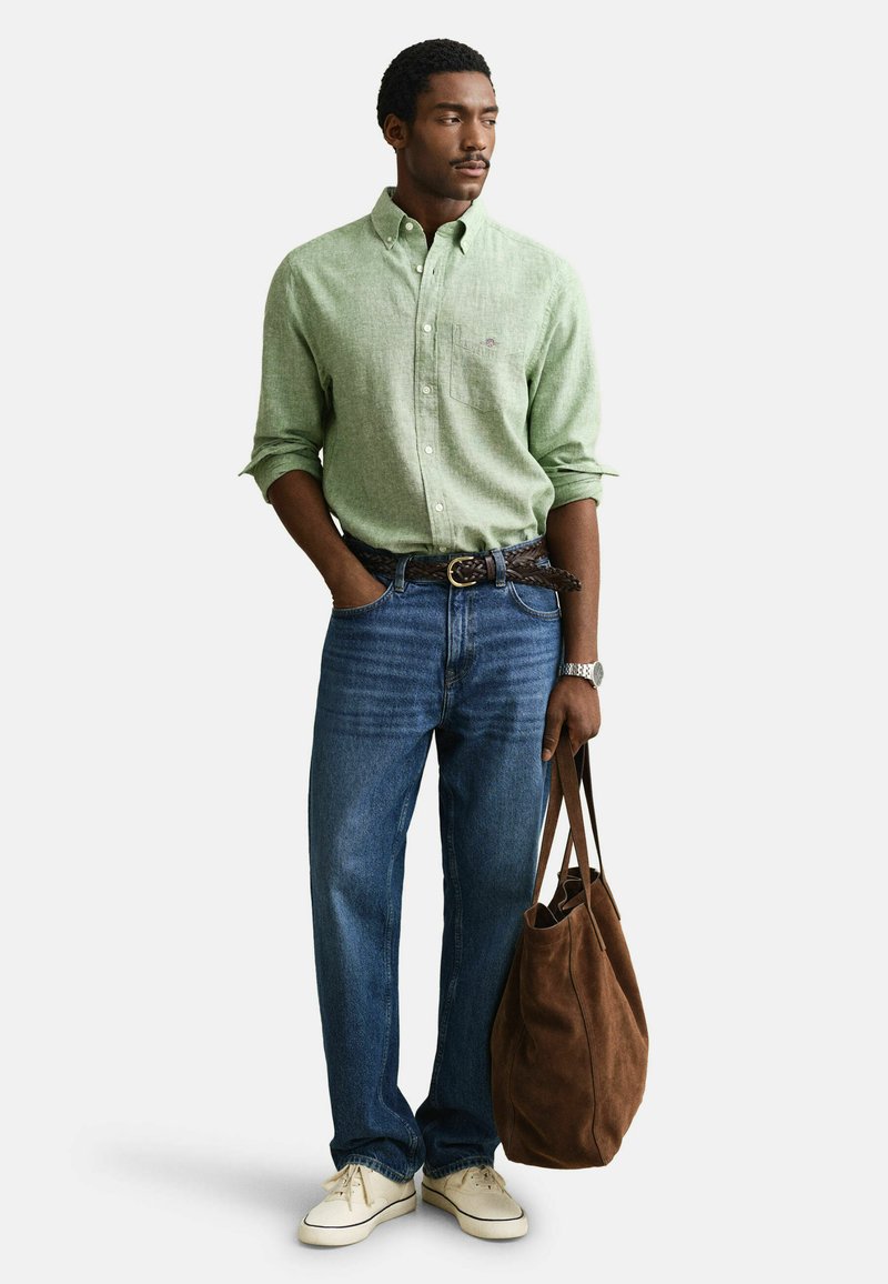 Hombre con camisa de botones verde claro, vaqueros azules, zapatillas blancas, sosteniendo un bolso tote marrón, de pie con una mano en el bolsillo.