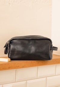 Zwarte leren toilettas met een rechthoekige vorm, ritssluiting, zijhandvat en gegraveerd logo aan de voorkant, geplaatst op een houten oppervlak.