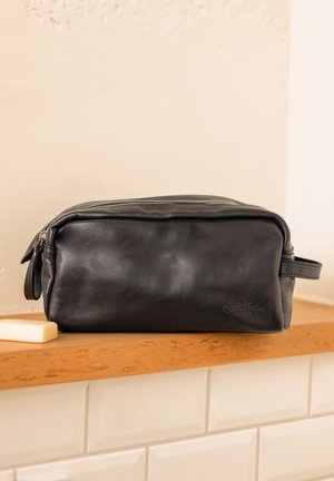 Schwarz Leder Kulturbeutel mit rechteckiger Form, Reißverschluss, seitlichem Griff und geprägt Logo auf der Vorderseite, platziert auf einer Holzoberfläche.