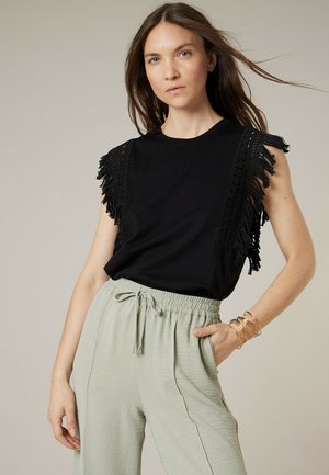 Femme en haut noir sans manches avec détails en franges et pantalon gris clair à cordon, posant avec une main dans la poche sur un fond neutre.