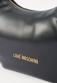 Bolsa de couro preto com uma forma curvada, detalhe em logotipo dourado "LOVE MOSCHINO" e fecho superior com zíper. Textura suave com um acabamento polido.