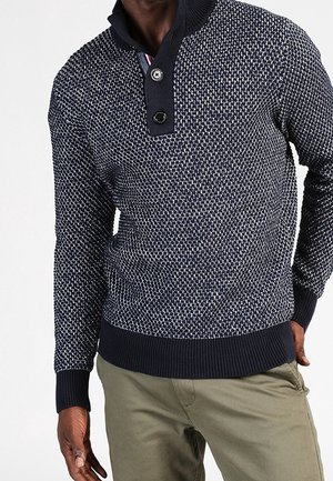 Mężczyzna w granatowo-białym swetrze z teksturowanym splotem, z dwoma guzikami na kołnierzu, w oliwkowozielonych spodniach.