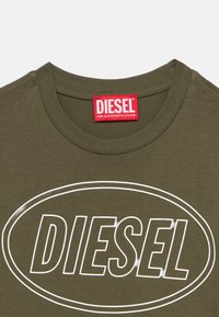 Ελιά πράσινο t-shirt Diesel με λευκό περιγραμμικό οβάλ λογότυπο και έντονο κείμενο "DIESEL" στο μπροστινό μέρος, και κόκκινη ετικέτα της μάρκας μέσα στον λαιμό.