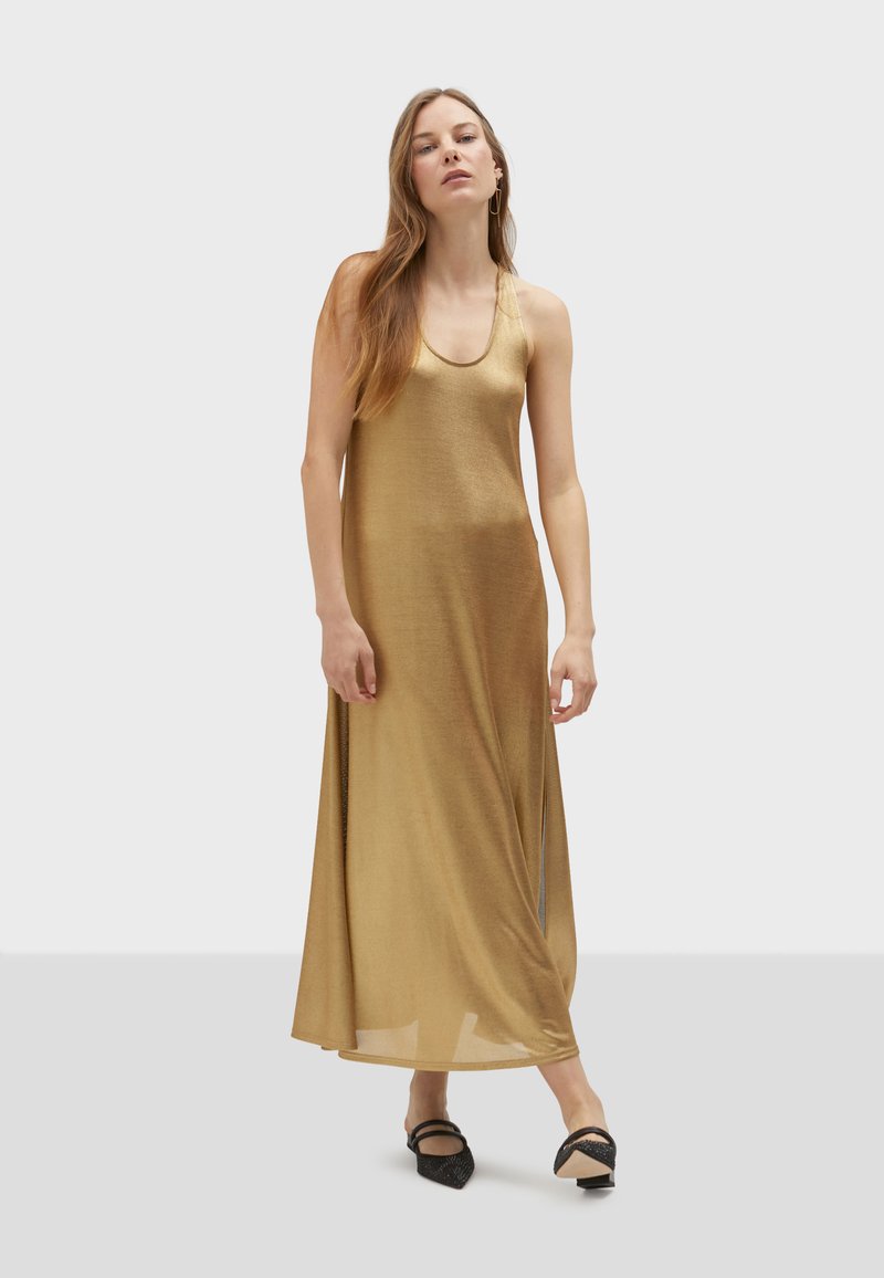 Zlaté, bezramínkové maxi šaty vyrobené z lehkého, lesklého materiálu; mají výstřih do vodorovna a vysoké boční rozparky. Na sobě má černé boty.