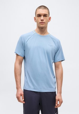 Jaunas vyras stovi, vilki šviesiai mėlyną trumpomis rankovėmis Ted Baker Sport marškinius ir tamsius šortus prieš paprastą šviesią foną.