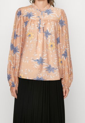 Kobieta ubrana w brzoskwiniową bluzkę w niebieskie i żółte kwiaty z długimi bufkami, połączoną z czarną plisowaną spódnicą.