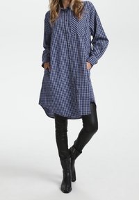 Blue and white checkered shirt dress with a buttoned front, long sleeves, and side pockets, paired with black fitted leggings and knee-high boots.